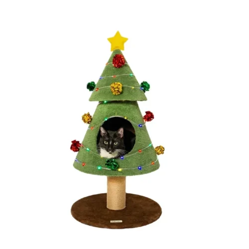A large black and white tuxedo cat peeking out from inside a festive green Christmas tree-shaped cat condo with colorful lights and pom-poms, featuring a sisal scratching post base.