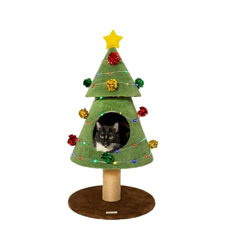 A large black and white tuxedo cat peeking out from inside a festive green Christmas tree-shaped cat condo with colorful lights and pom-poms, featuring a sisal scratching post base.